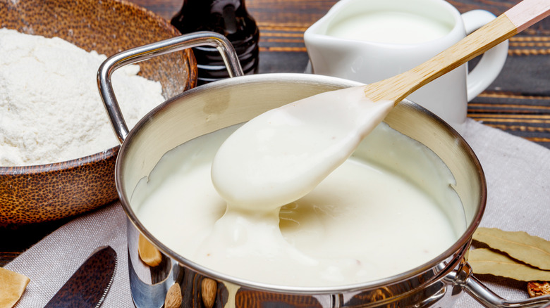 Creamy béchamel sauce in pan with wooden spoon on a placemat with ingredients in background.