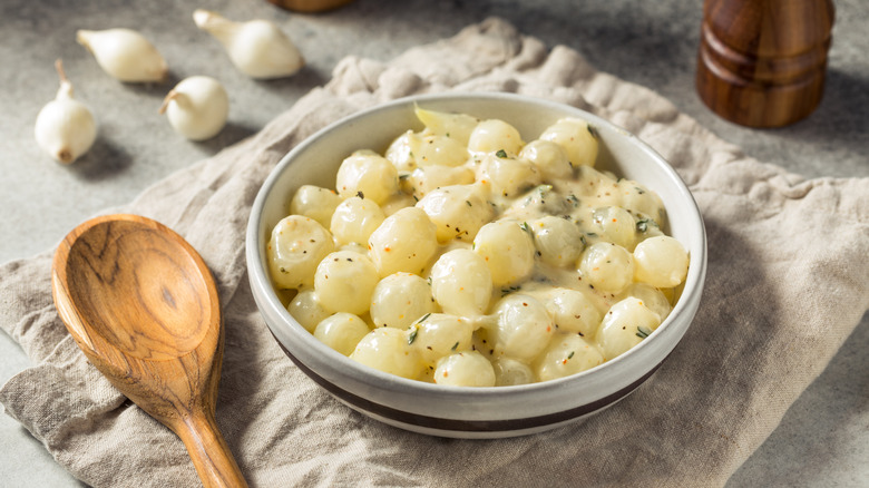 Small bowl of creamed pearl onions with thyme and wooden spoon on cloth mat.