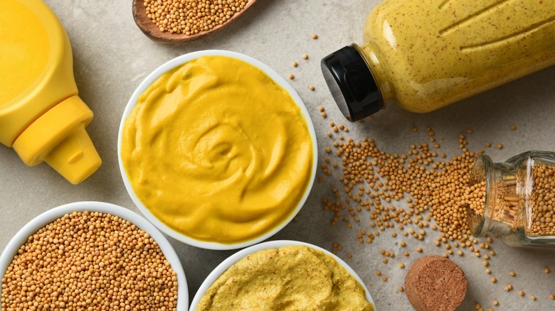 Yellow and brown prepared mustard and mustard seeds in bottles and bowls.