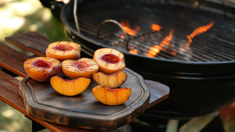 A tray of grilled peach halves beside a charcoal grill.