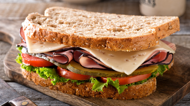 A fresh ham sandwich made with wheat bread (plus pickles, cheese, tomatoes, and lettuce) on a wood cutting board