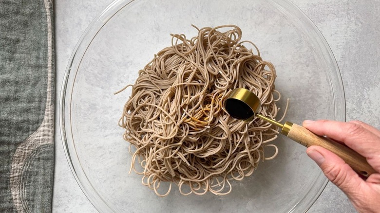 Adding soy sauce to cooked soba noodles in large glass bowl