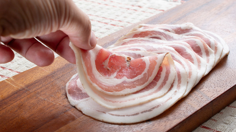 A person's hand is seen removing a slice of pancetta from a cutting board.