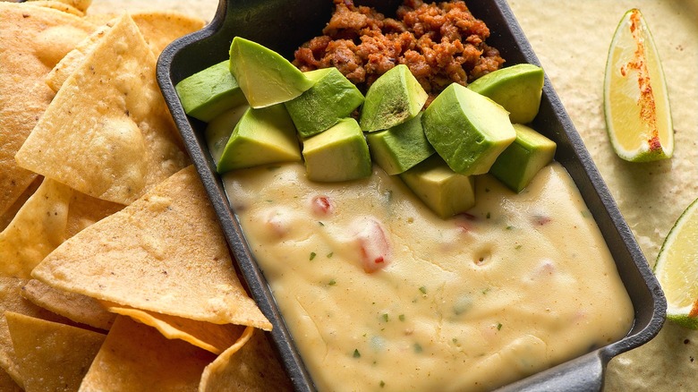 Rectangular metal pan filled with warm queso, ground meet, and chopped avocado surrounded by tortilla chips and lime wedges