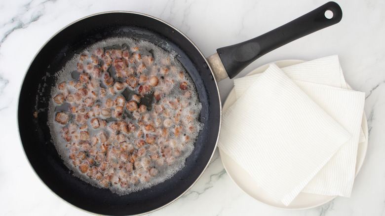 crispy bacon bits cooking in skillet next to lined plate