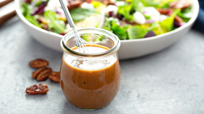 Dark salad dressing in a small glass with a mini whisk in front of a shallow bowl of salad