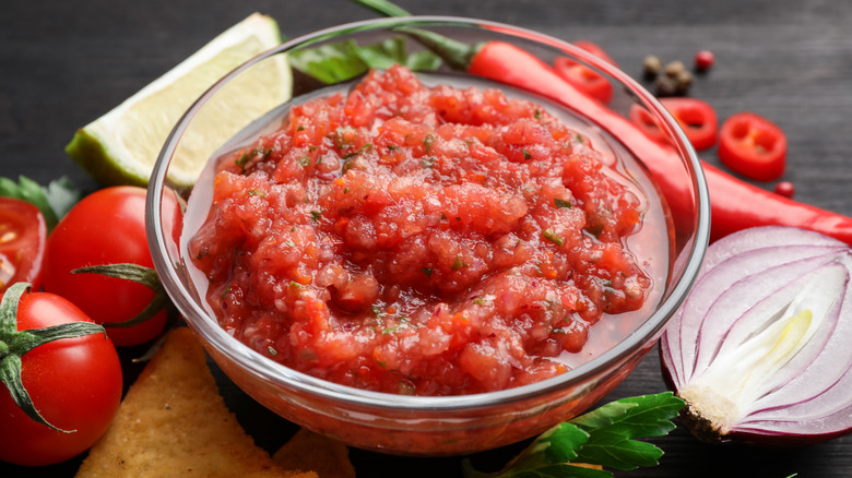 Spicy salsa sauce in a clear glass bowl surrounded by nachos and ingredients on table, closeup.