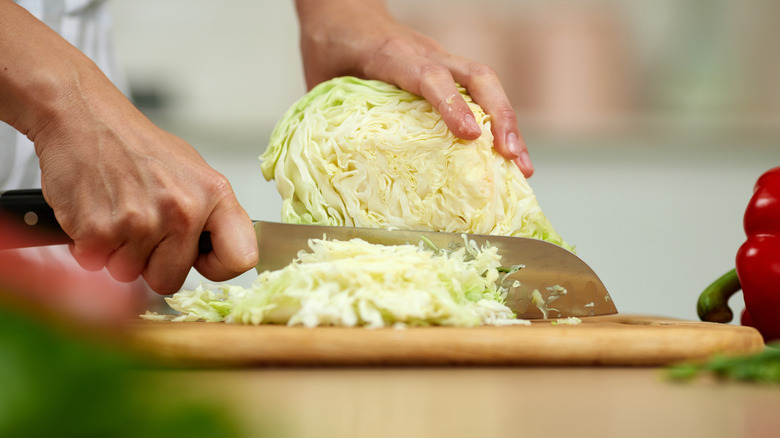Person's hands using a knife to finely chop fresh green cabbage on a wooden board, preparing healthy food in a kitchen.