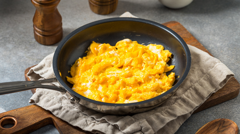 A stainless steel pan of scrambled eggs sits on a dish towel on a wooden cutting board