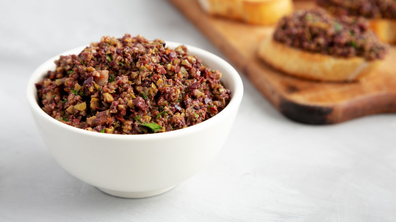 A white bowl of tapenade with tapenade-topped appetizers in the background out of focus