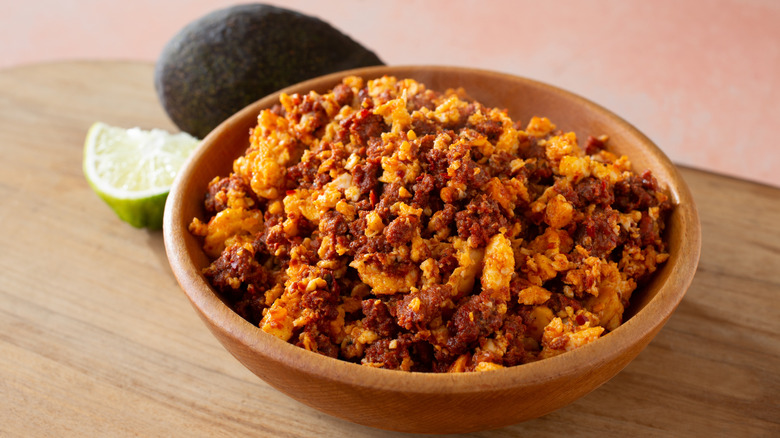 Chorizo scrambled eggs in a wooden bowl with avocado and a lime wedge in the background