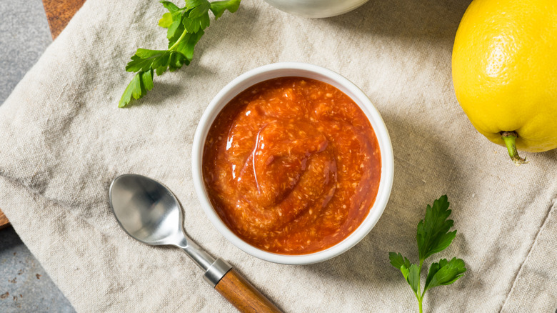 A small bowl of cocktail sauce is arrranged in a linen napkin near a spoon, herbs, and a lemon.