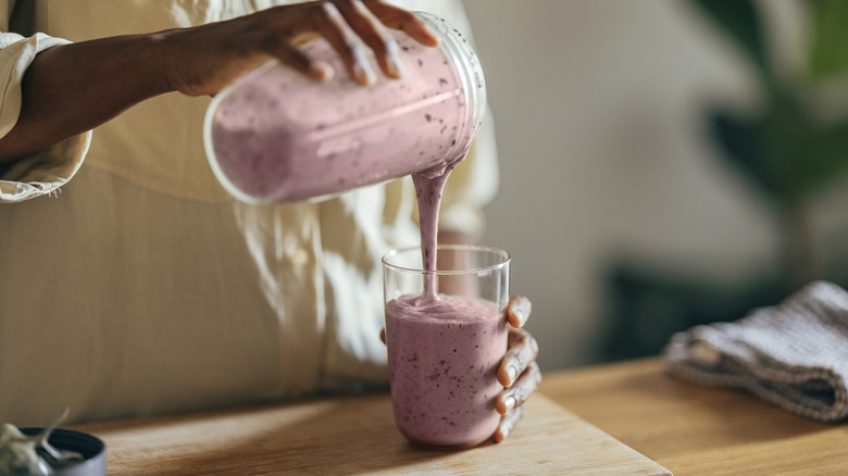Close-up of hands of a person wearing beige shirt and pouring smoothie into a glass on a kitchen table