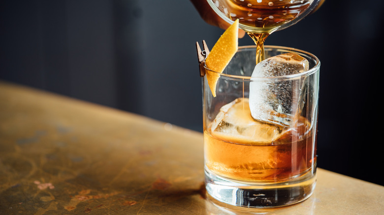 An old fashioned cocktail being poured into a glass with ice and orange peel