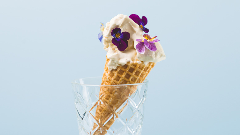 An ice cream cone balancing in a glass topped with vanilla ice cream and purple flowers