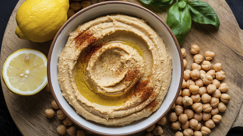 Bowl of hummus on wooden board next to chickpeas, lemon, and basil.