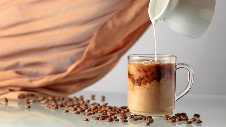 Milk is poured into a glass cup of coffee from a pitcher.