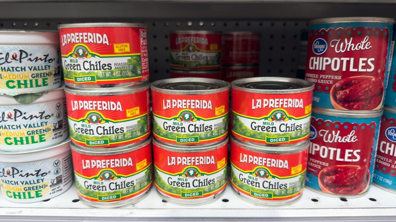 Rows of chopped canned green chiles at the supermarket, a stack of canned whole chipotle peppers next to them.