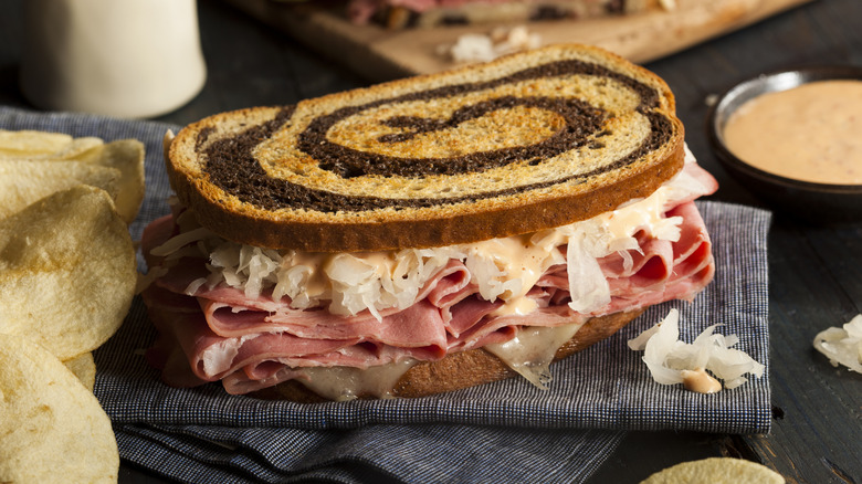 A Reuben sandwich on a cloth napkin surrounded by potato chips and dipping sauce