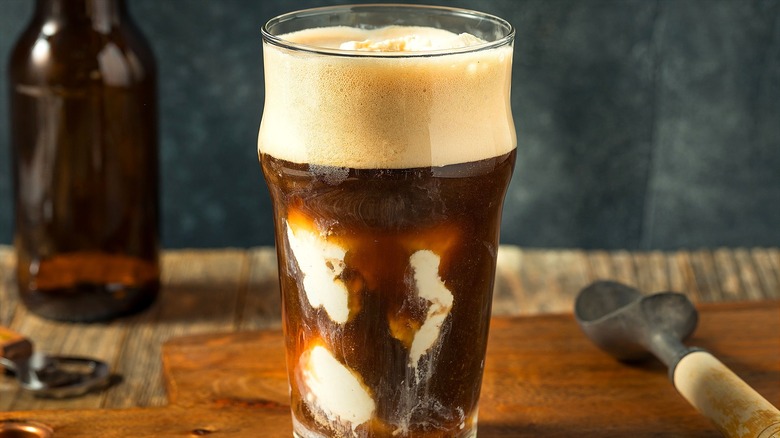 Homemade root beer float in a clear glass on a wooden serving board next to an ice cream scoop