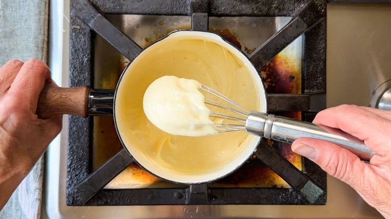 Thick white cheese sauce on whisk over small sauce pan on stovetop
