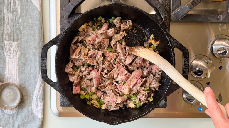 Browning shaved beef with peppers and onions in cast iron skillet with wooden spoon