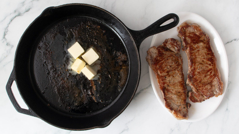 chunks of butter in a black frying pan next to cooked steaks on a white plate