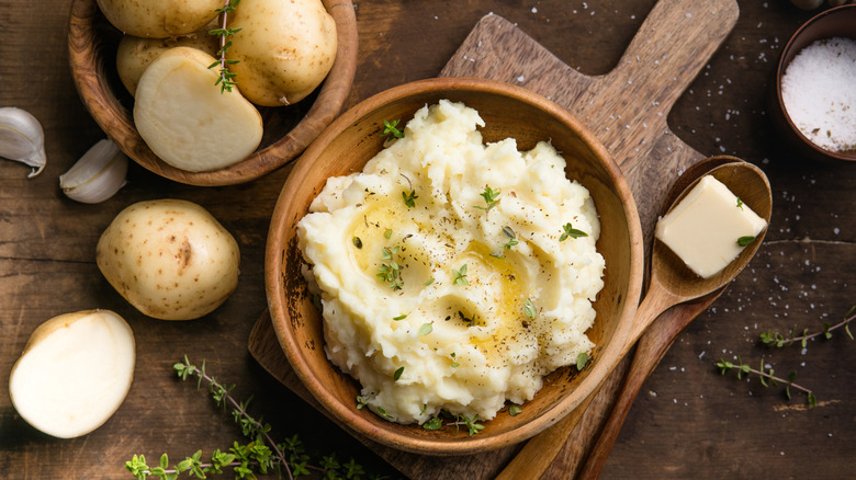 Mashed potatoes topped with butter and herbs in a wooden bowl on a wooden cutting board