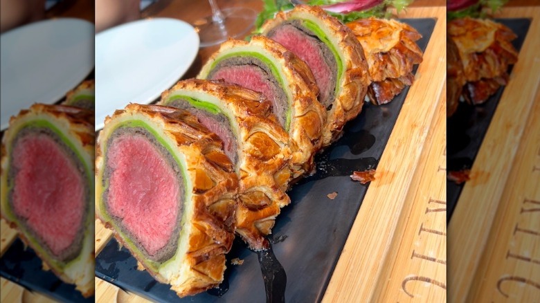 Slices of Gordon Ramsay's famous beef Wellington, consisting of medium-rare beef filets wrapped in prosciutto, mushroom paste, and puff pastry, lay on a board atop a table, with a plate, silverware, and glasses of wine and water in the background