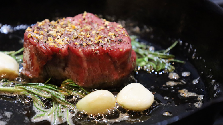 Filet mignon seasoned with salt and pepper in cast iron skillet next to garlic and rosemary
