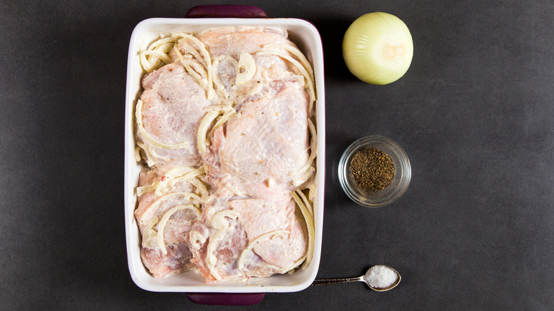 chicken in a baking dish with onions and mayonnaise marinade. Black surface in background with raw onion, jar of pepper, spoonful of salt