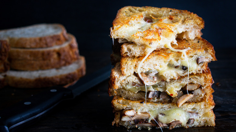 A stack of grilled cheese sandwiches with cheese and mushrooms in them and bread slices in the background.