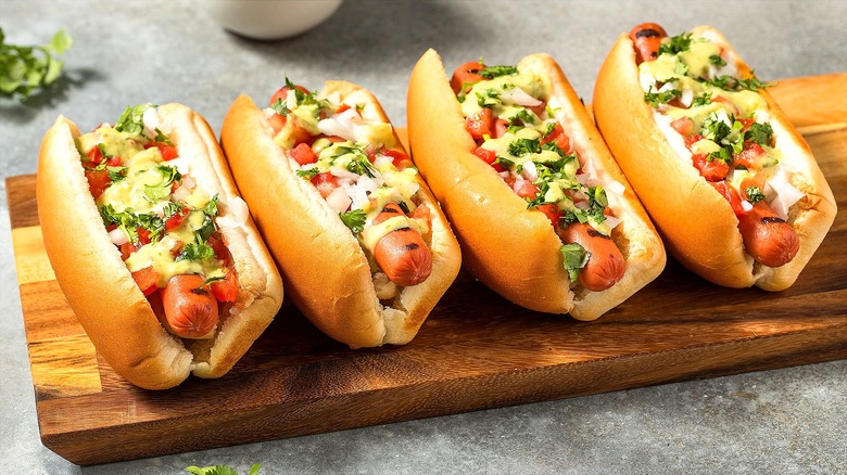 Four hot dogs with salsa, avocado cream sauce, and cilantro on a wooden serving board