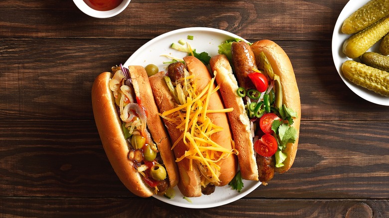 Three hot dogs with various toppings like olives, shredded cheese, cilantro, and cherry tomatoes on a white plate