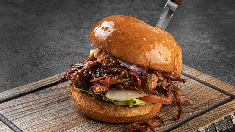 A pulled pork sandwich with a steak knife down the center securing it to a cutting board.