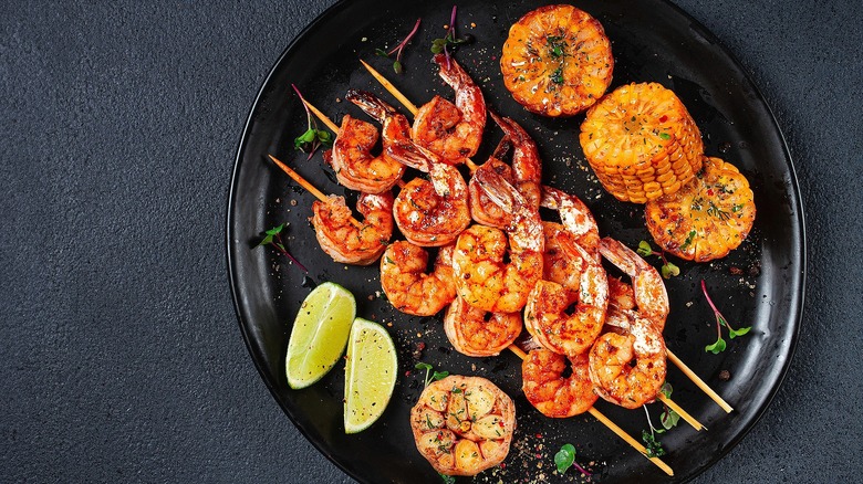 Top-down view of seasoned shrimp skewers, corn, garlic, and lime wedges on a black plate.