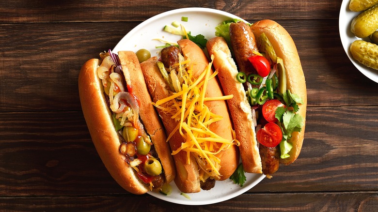 Three hot dogs with shredded cheese, olives, and tomatoes on a white plate.