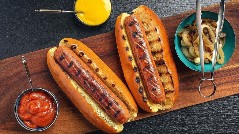 Two grilled hot dogs in buns on a board with bowls of ketchup, mustard, and caramelized onions.