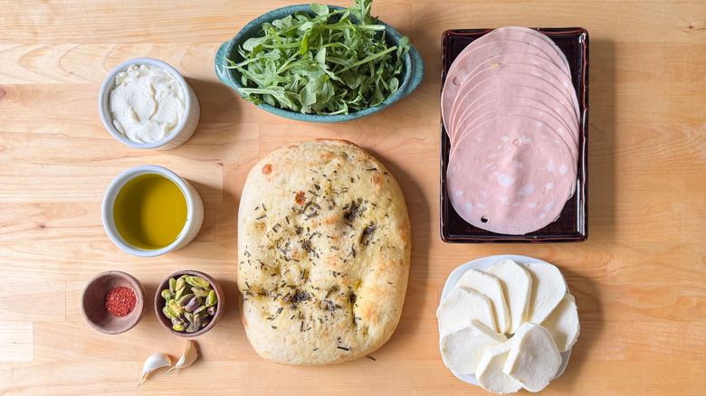 Loaf of focaccia, mortadella, arugula, cheeses, pistachios, olive oil, chile flakes, and garlic cloves on wooden surface