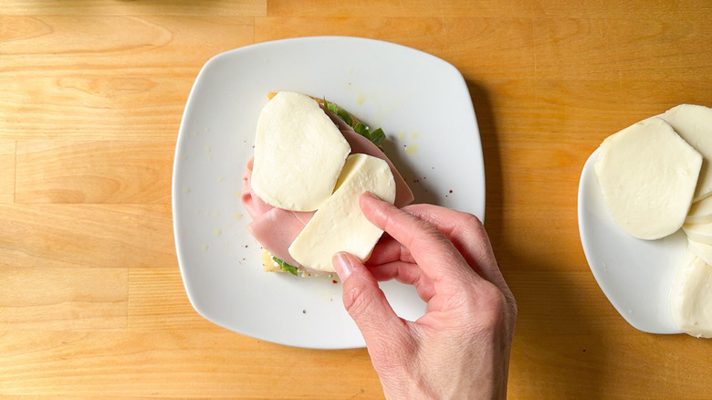 Hand placing fresh mozzarella slices on top of mortadella on focaccia sandwich on plate