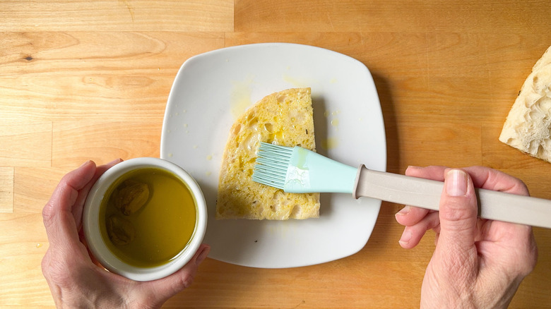 Hand brushing garlic oil on focaccia slice on plate