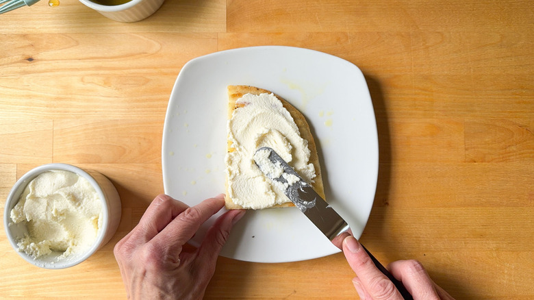 Hand spreading ricotta cheese on slice of focaccia on plate