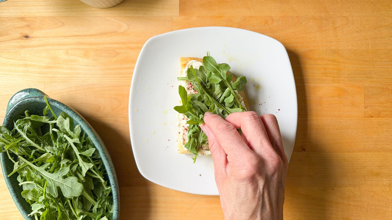Hand putting arugula leaves on focaccia bread slice with ricotta and chile flakes on plate