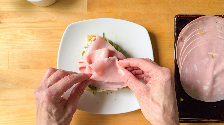 Hand placing mortadella slices on focaccia sandwich on plate
