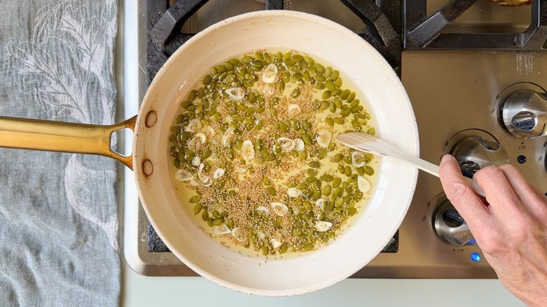 Stirring garlic slivers, pepitas, and sesame seeds in avocado oil in skillet on stovetop with wooden spoon