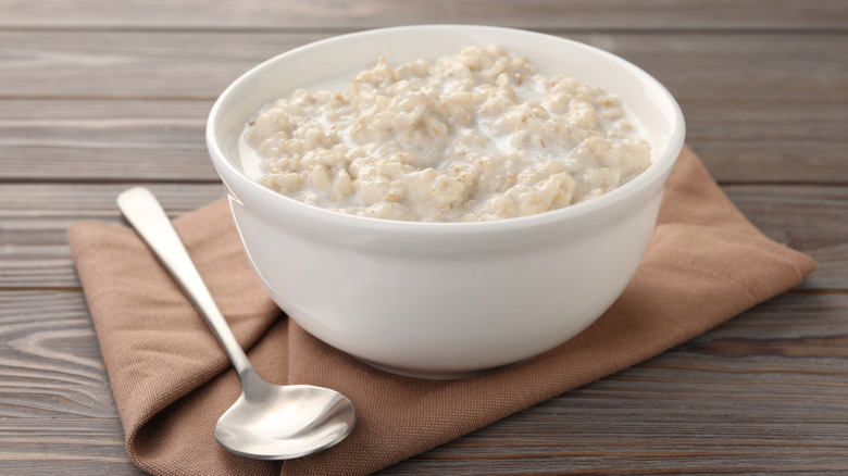 A bowl of plain oatmeal on a cloth on a wooden table with a spoon alongside it.