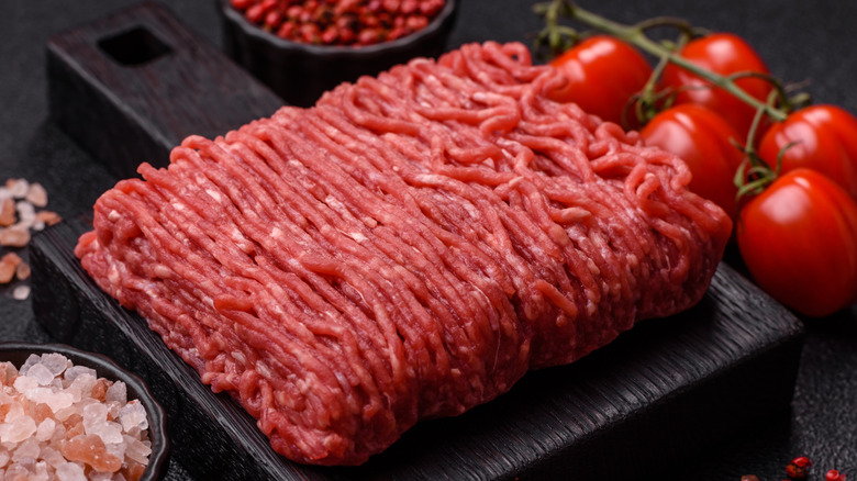 Raw ground beef on thick cutting board with salt and tomatoes nearby.