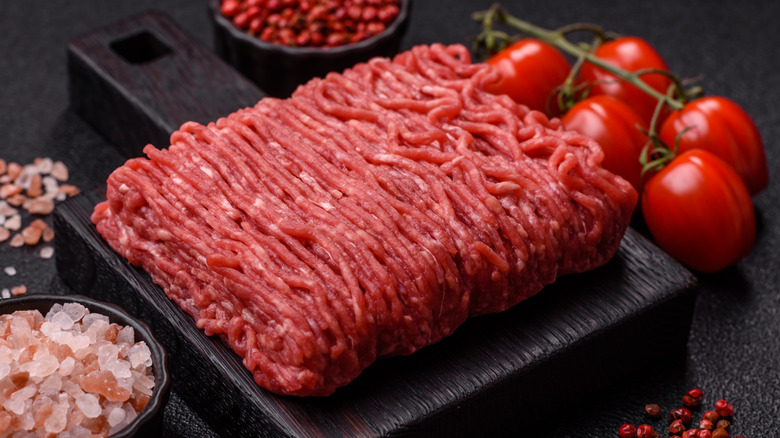 A block of ground beef is on a black cutting board arranged near tomatoes and salt.