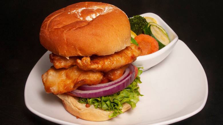 A fried grouper sandwich with onion and tomato served with grilled veggies