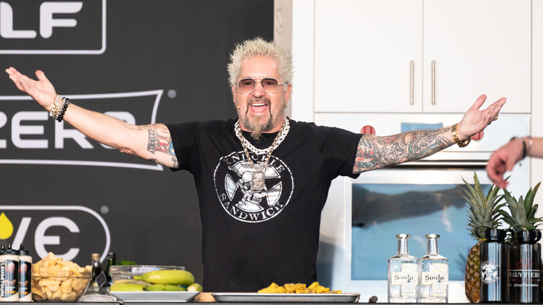 Guy Fieri smiling with his arms extended during a cooking demonstration